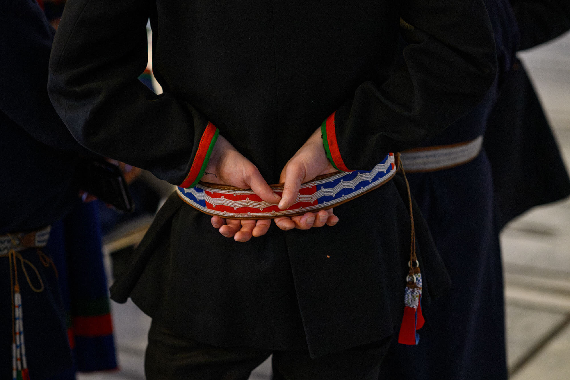 Nærbilde av ryggen til en mann i mørk kofte med belte i samiske farger.